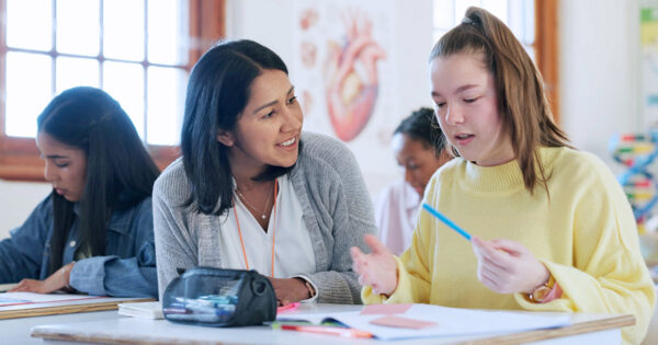 A teacher and student talk through classwork together at a classroom desk.